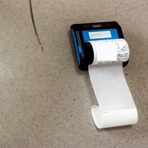 Petite imprimante thermique noire et bleue posée au sol, une longue étiquette blanche sortant de l'appareil